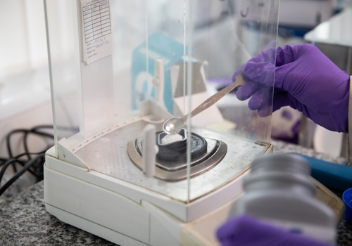 A person wearing purple gloves placing a white powder onto a digital lab balance using a spatula.